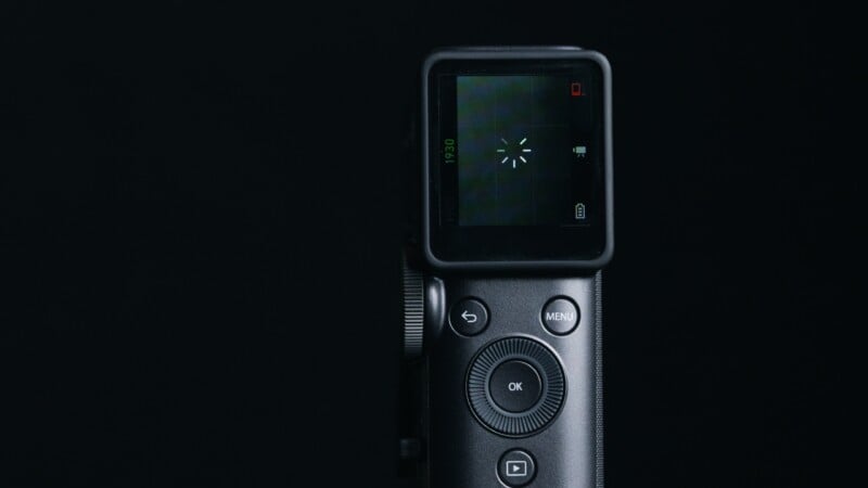 A close-up of a digital camera's rear screen and control buttons, displaying a loading symbol on the screen, against a black background.