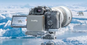 A professional camera covered in frost sits on a tripod in an icy landscape, filming a polar bear walking across snow and ice, with more icebergs and water in the background.