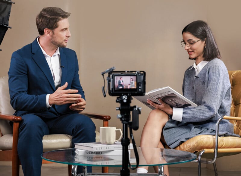 Un hombre con traje azul y una mujer con gafas y un suéter gris se sientan en sillones y conversan. Una cámara montada en un trípode los grabó. La mujer sostiene una revista abierta; Los libros y las tazas se colocan sobre la mesa de cristal de enfrente.