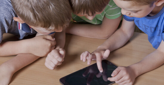 Three young boys closely gathered around a tablet on a wooden table, interacting with the touchscreen together and focusing intently on the device.