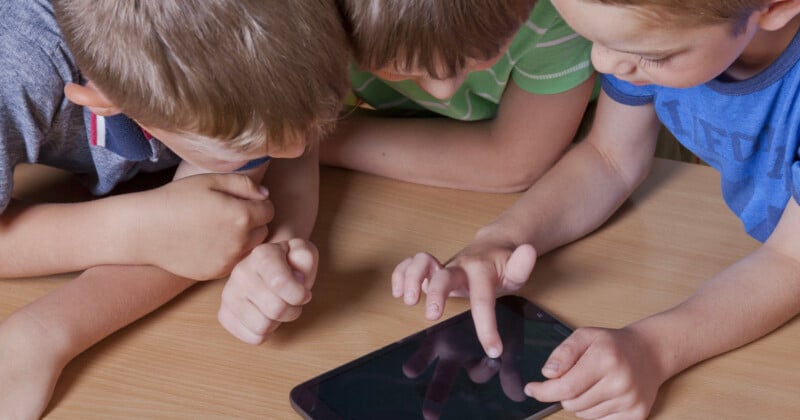 Tres niños pequeños se sientan juntos alrededor de una tableta sobre una mesa de madera, interactúan juntos con la pantalla táctil y se concentran en el dispositivo.