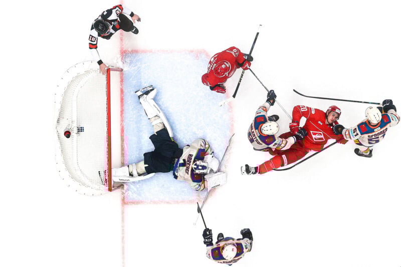Aerial view of hockey players in action near the goal net. The goalie is sprawled on the ice while players from both teams crowd around, competing for control of the puck. The ice and goal area are clearly visible.