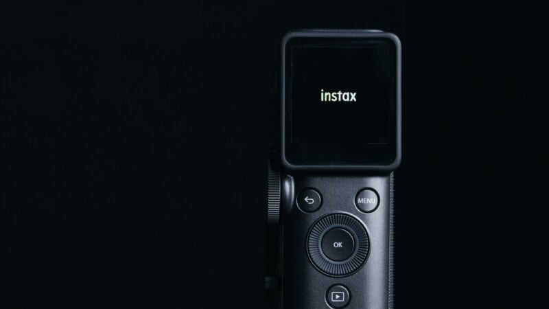 A close-up of a black digital camera against a dark background, with the screen displaying the word "instax" in white text.