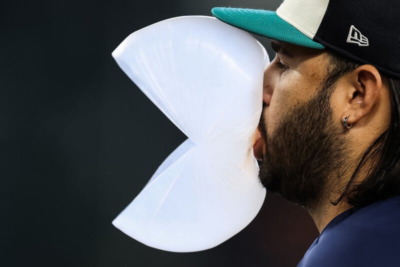 A baseball player in a cap blows a large bubble with chewing gum, captured mid-pop with the bubble split open in a V shape near his face.