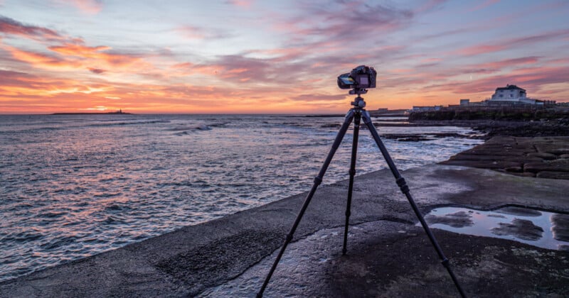 A camera on a tripod is set up on a concrete pier, facing a colorful sunset over the ocean, with a house and lighthouse visible in the distance. The sky is painted with shades of pink, orange, and purple.