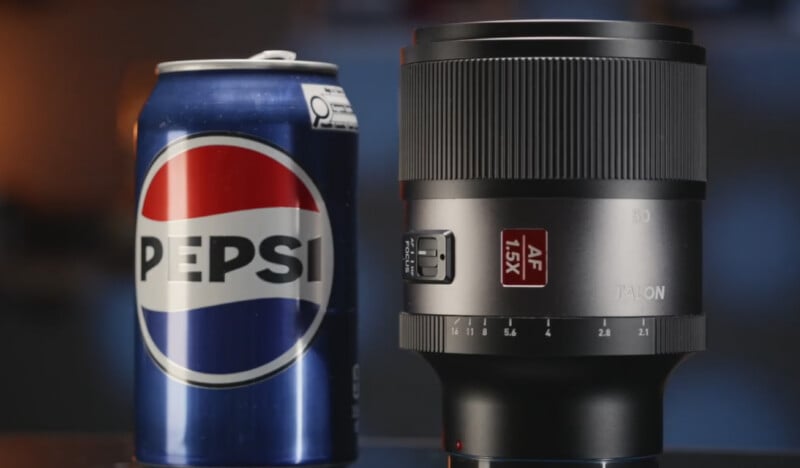A Pepsi can stands next to a TALON camera lens with an AF 1.5X label, both positioned upright on a blurred background surface.