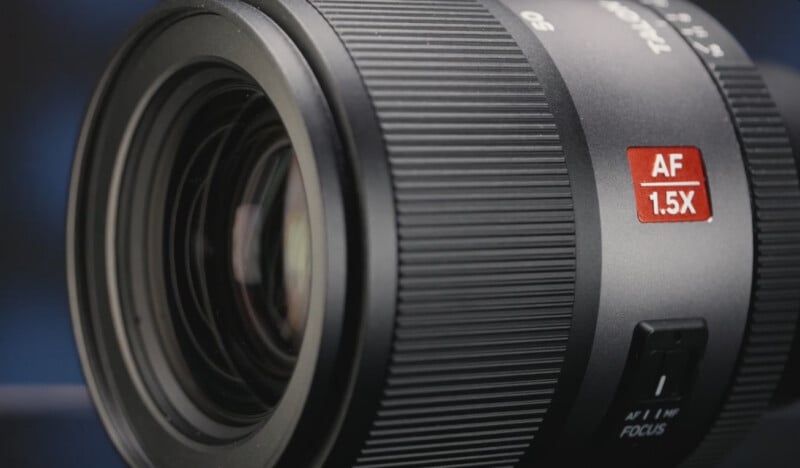 Close-up of a camera lens with a textured focus ring, displaying a red label marked "AF 1.5X" and a switch labeled "AF/MF FOCUS" on the side. The background is blurred and dark.