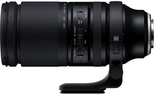 A black telephoto camera lens with zoom and focus rings, multiple control switches, and a tripod mounting collar, shown in a side view against a white background.