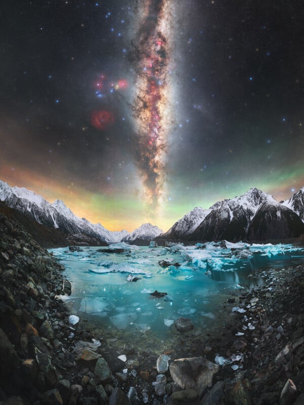 A vivid night sky with the Milky Way stretches over snow-capped mountains and a rocky, icy lake. The colorful galaxy is reflected in the still turquoise-blue water amid scattered rocks and ice.