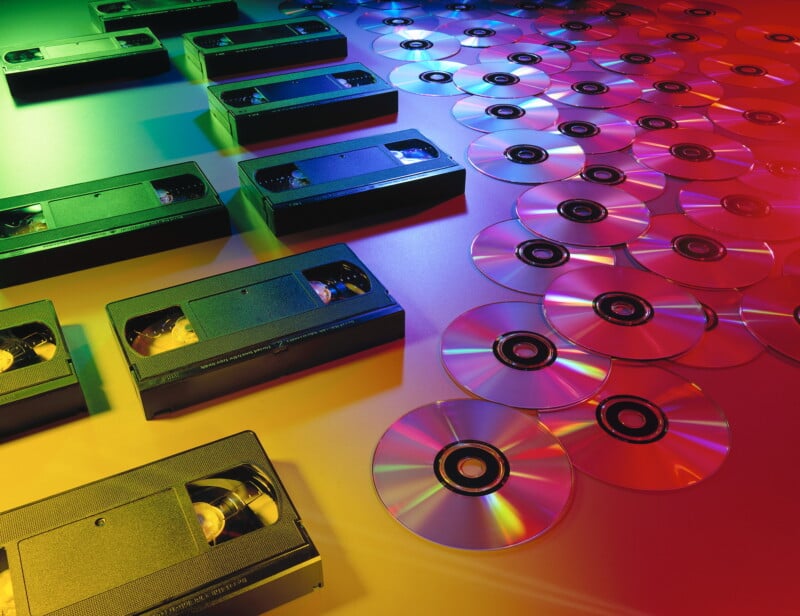 Several VHS tapes and numerous compact discs (CDs or DVDs) are spread out on a surface. The scene is lit with colorful, gradient lighting in green, yellow, and red tones, creating a vibrant, retro feel.