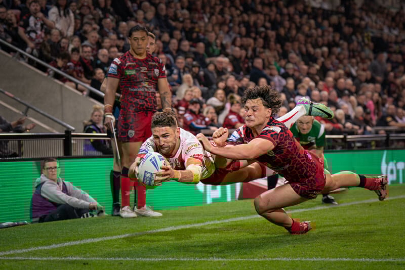 A rugby player in white dives to score a try while a player in red attempts to tackle him. The stadium is packed with spectators watching the intense moment near the sideline.