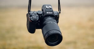 A Nikon Z series digital camera with a zoom lens hangs from a black strap, suspended in front of a blurred, beige outdoor background.