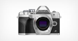 A silver and black Olympus OM-D digital camera body is shown facing forward on a plain white background, without a lens attached. The camera features textured grip and classic retro styling.