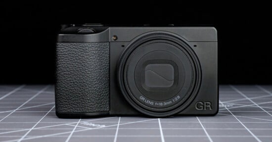 A compact black digital camera with a textured grip and a closed lens cover sits on a gray grid-patterned surface. The letters "GR" are visible on the bottom right of the camera's front.