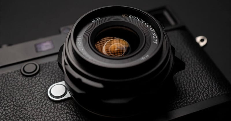 Close-up of a black camera with a textured body, focusing on the lens which has "Epoch Coating 73" written on the rim. The background is dark and the lens reflects a soft light.