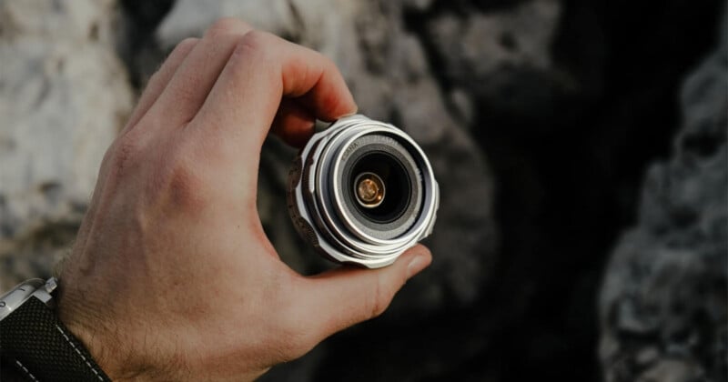 A hand holding a small, round camera lens or optical device against a blurred rocky background.