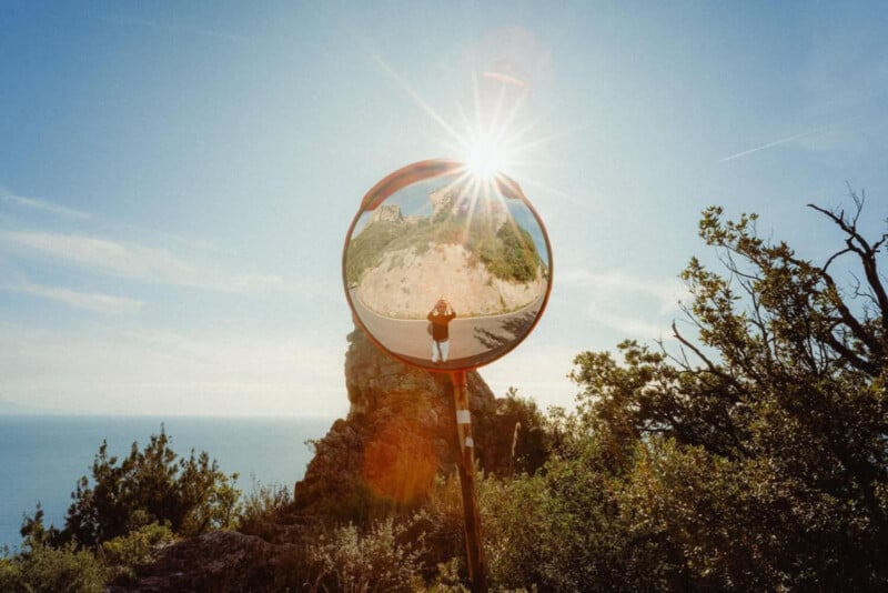 A round convex mirror outdoors reflects a person standing on a rocky path, with sunlight shining above and a scenic, green landscape in the background. Bushes frame the scene under a clear blue sky.