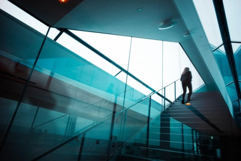 A person walks up a modern glass staircase in a building with large glass windows and blue-tinted reflections, creating a sleek, contemporary atmosphere.