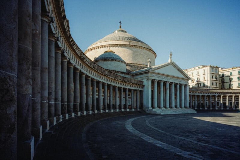 A wide plaza with curved colonnades and a domed neoclassical building under a clear blue sky, with statues on the roof and surrounding city buildings in the background.