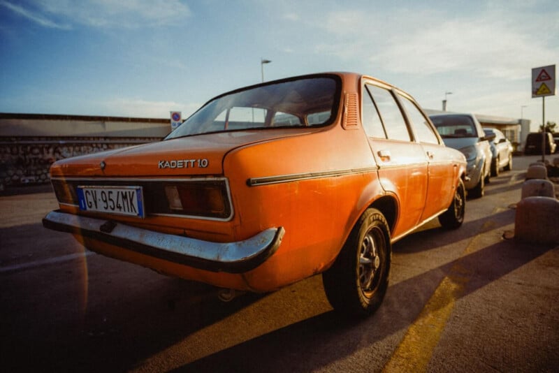 A vintage orange Opel Kadett 1.0 car is parked on a sunlit street next to another vehicle, with signs and buildings visible in the background.