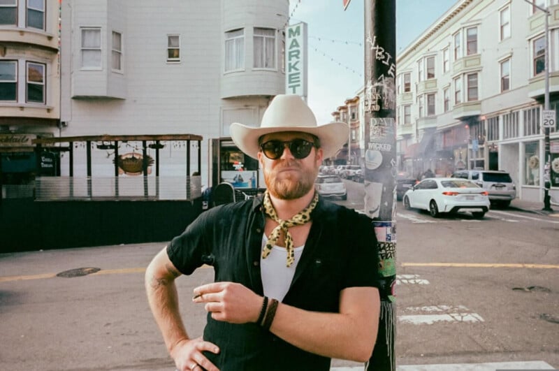 A man wearing a cowboy hat, sunglasses, a black shirt, and a yellow scarf stands by a pole on a city street, holding a cigarette. Buildings, cars, and street signs are visible in the background.