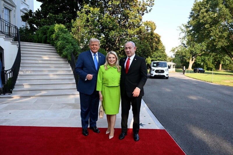 Three people stand on a red carpet outside a large white building, with trees and a van in the background. They are dressed formally, smiling at the camera, with one woman in a bright green outfit between two men in suits.