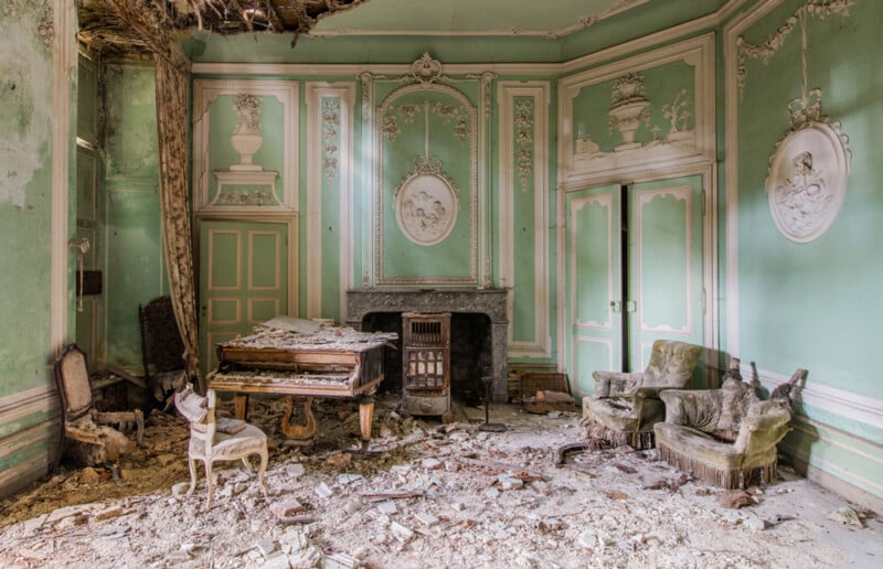 An abandoned, ornate room with pale green walls, white molding, a damaged piano, old armchairs, debris on the floor, and a hole in the ceiling letting in light. The space appears dilapidated but once elegant.