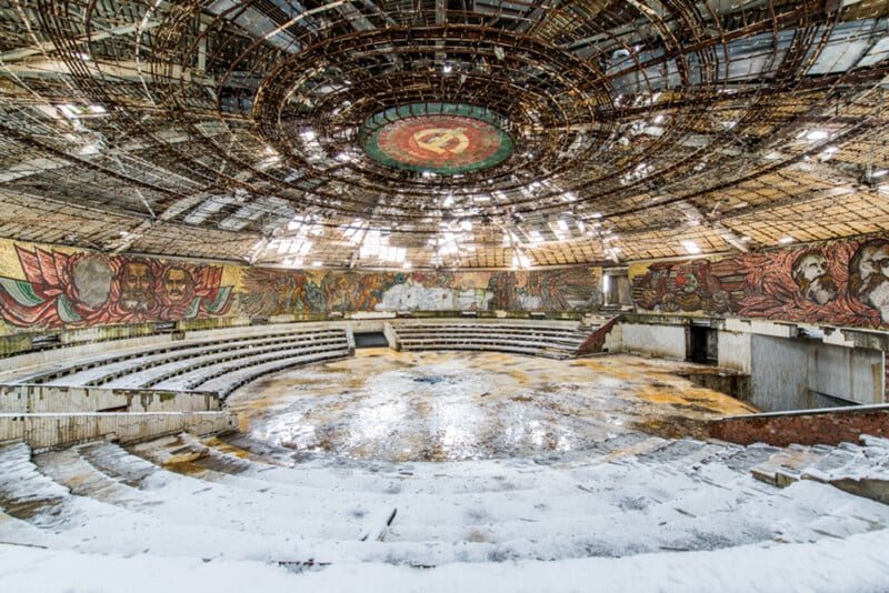An abandoned, circular amphitheater with snow covering the floor, rusted metal roof structure, and colorful murals of faces and scenes on the walls. A faded red emblem is visible in the center of the ceiling.