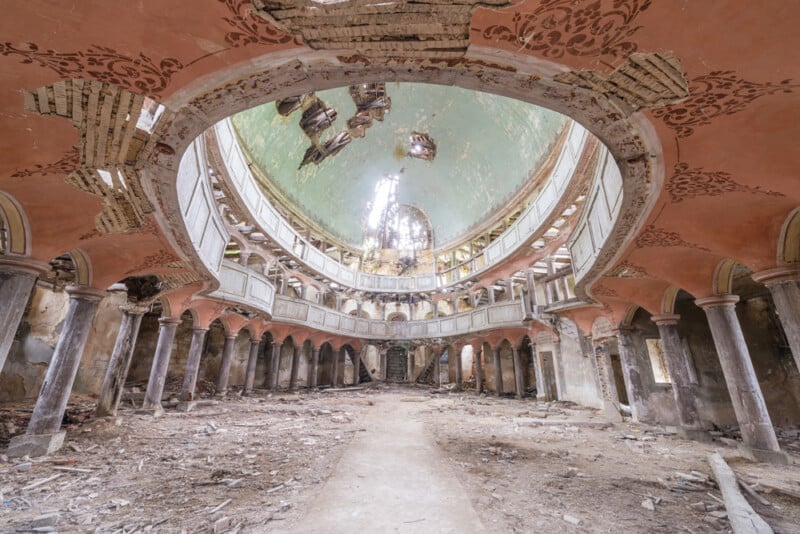 An abandoned, decaying church interior with a large dome ceiling, peeling paint, broken columns, and debris scattered across the dusty floor. Sunlight streams through holes in the roof, illuminating the derelict space.