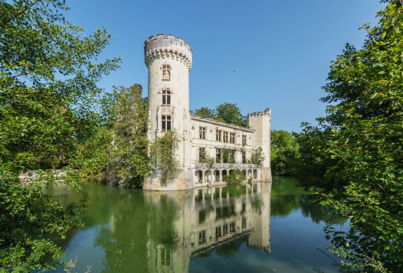 A white stone castle with a round tower stands surrounded by trees, reflected in the still green water of a moat, under a clear blue sky.