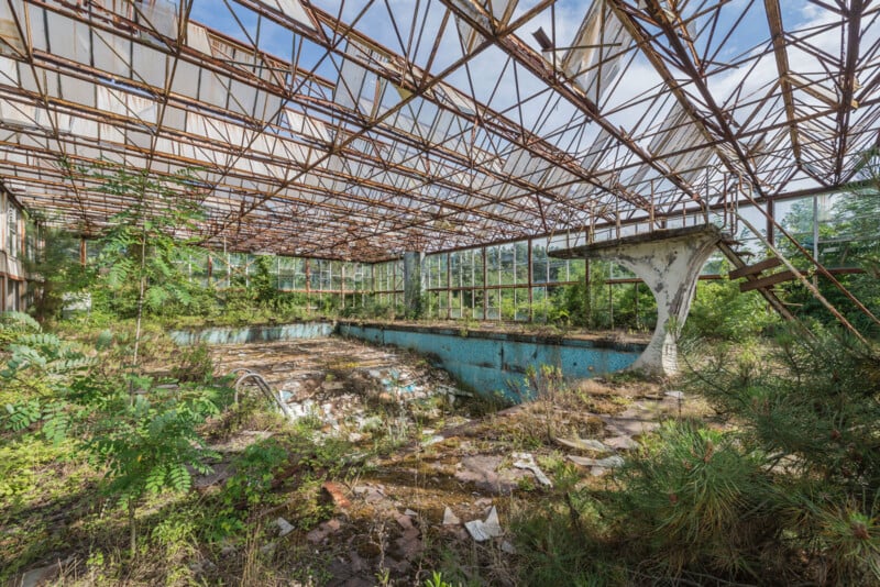 An abandoned indoor swimming pool overtaken by vegetation, with a rusted, partially collapsed roof, broken tiles, and debris scattered around the empty, dirty pool. Sunlight filters through the open ceiling.