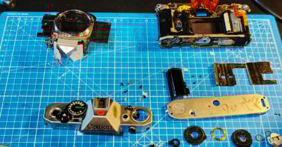 Disassembled Canon AE-1 film camera parts spread on a blue grid cutting mat, including the body, top plate, screws, dials, and other components. Tools and small pieces are visible around the workspace.