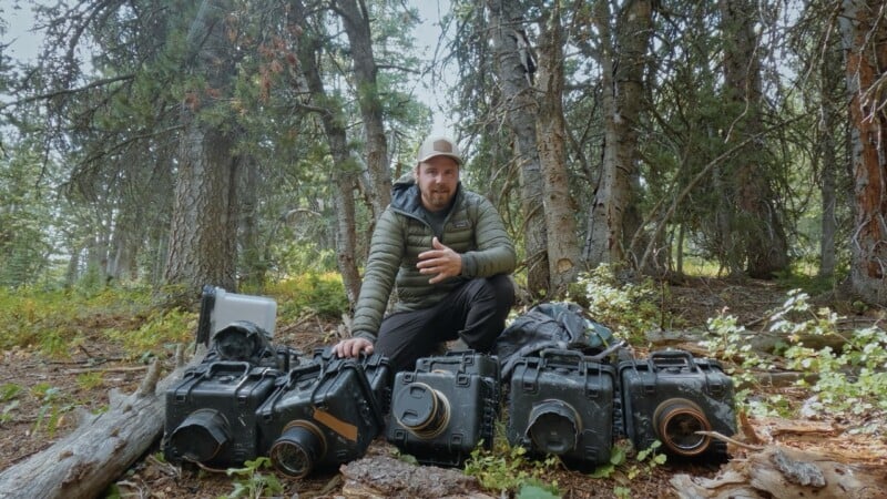 Un hombre vestido con ropa de exterior está sentado en un claro del bosque, rodeado por varias cajas duras negras y resistentes. La luz del sol se filtra a través de los altos árboles del fondo. Parecía estar hablando o gesticulando.