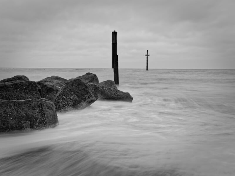 Black and white photo of a rocky shoreline with waves washing over the rocks. Two vertical posts stand in the water, one close to the rocks and one farther out. The sky is cloudy and overcast.