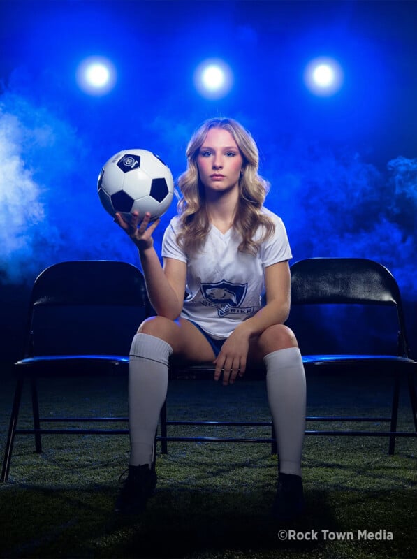 Una joven jugadora de fútbol con uniforme blanco está sentada en el banco, sosteniendo una pelota de fútbol en una mano y mirando con confianza a la cámara en un espectacular ambiente de luz azul. "Medios de comunicación de la ciudad del rock" La marca de agua es visible.