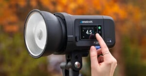 A hand adjusts the settings on a Westcott FJ200 strobe light, which is mounted on a tripod. The background is out of focus with autumn-colored foliage.