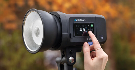 A hand adjusts the settings on a Westcott FJ200 strobe light, which is mounted on a tripod. The background is out of focus with autumn-colored foliage.