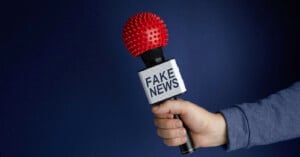 A hand holds a microphone labeled “FAKE NEWS.” The microphone’s head is covered with a red, spiky ball resembling a virus, against a plain dark blue background.