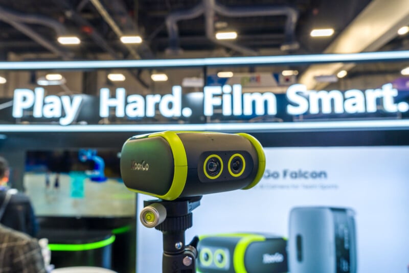 En la exposición se exhibe un equipo de cámara verde y negro montado en un trípode. Detrás hay un cartel que dice "jugar duro. Película inteligente." Más imágenes de productos y personas en el fondo borrosas.