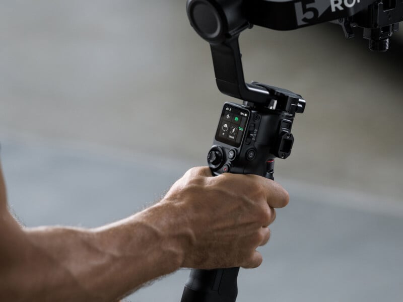 A close-up of a person's hand holding and operating a professional camera gimbal stabilizer with a display screen and control buttons, against a blurred background.