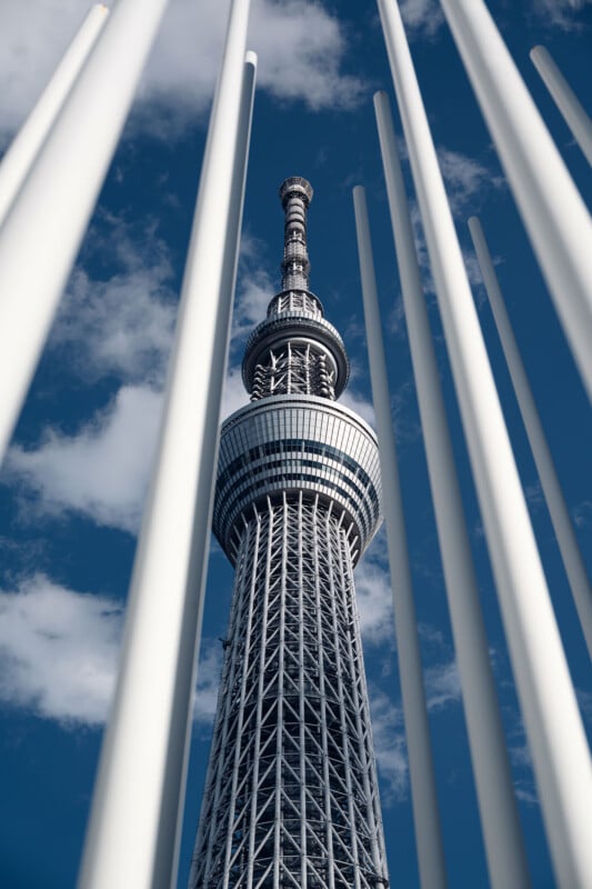 El Tokyo Skytree se alza sobre un fondo de cielo azul y nubes blancas, con varios postes verticales blancos enmarcando el primer plano.