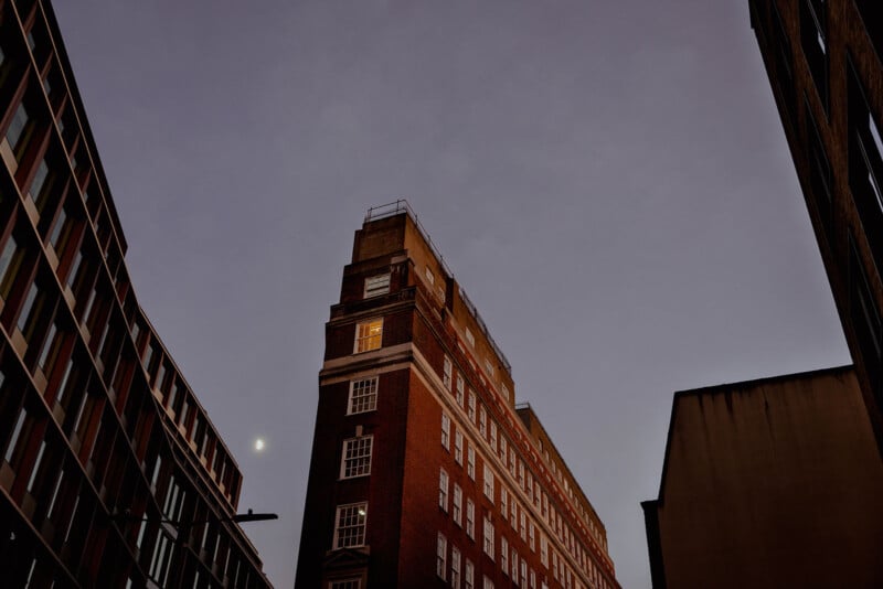 Al anochecer, un alto edificio de ladrillo se alzaba entre otros dos edificios, con las ventanas brillantemente iluminadas. El cielo es de color púrpura oscuro, con una luna pequeña y brillante visible cerca de la parte superior izquierda.