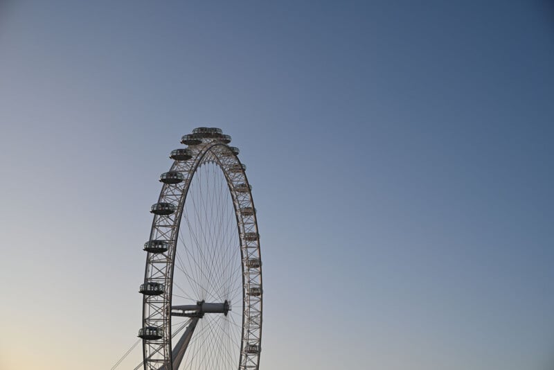 Una noria gigante se muestra parcialmente contra un cielo azul claro, con varias cabinas de pasajeros visibles a lo largo de su borde. Esta foto fue tomada desde abajo, resaltando la curvatura y altura de la estructura.