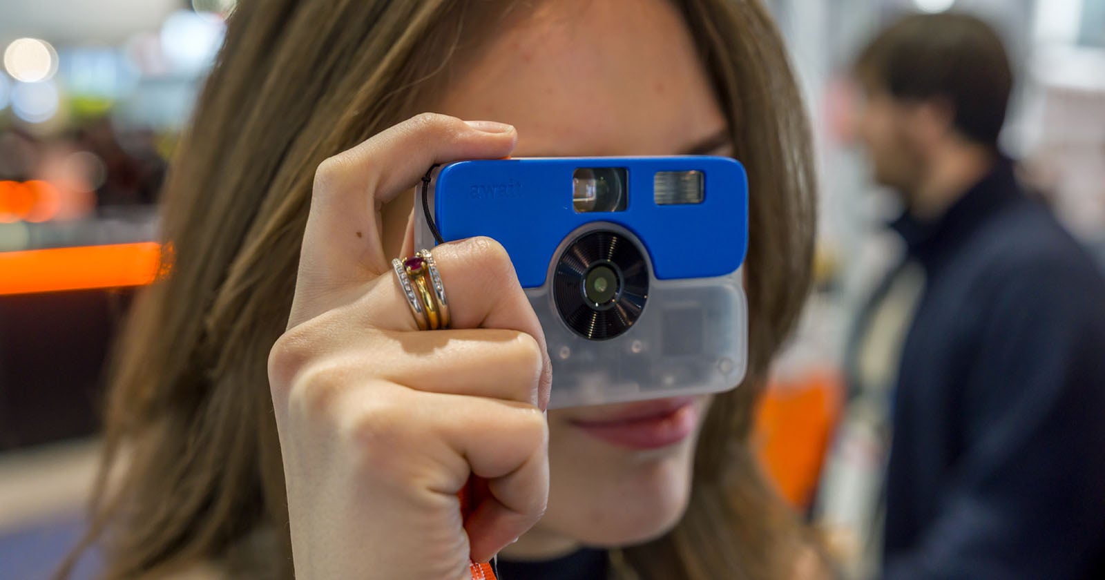 A person holds a blue and clear plastic camera up to their face, preparing to take a photo. The focus is on the camera and their hand, while the background is blurred.