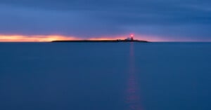 A distant lighthouse glows on a small island at dusk, its red light reflecting across calm blue water under a cloudy sky, with a thin strip of orange light on the horizon.