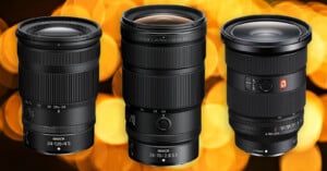 Three professional camera zoom lenses are shown standing upright side by side against a backdrop of large, out-of-focus golden lights. Each lens has visible brand and model markings.