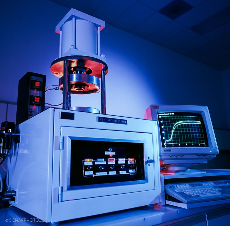 A laboratory machine labeled “BROOKFIELD 100” is positioned beside an old CRT computer monitor displaying a green and yellow graph. The setup is illuminated with blue and pink lighting in a lab setting.