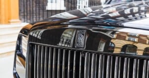 Close-up of the front grille and emblem of a black Rolls-Royce car, with reflections of nearby buildings visible on the shiny surface.