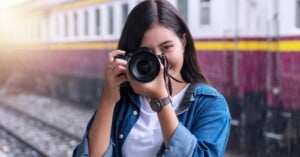 A young woman with long dark hair, wearing a blue denim jacket and white shirt, smiles while holding a camera up to her face at a train station, with train tracks and a train in the background.