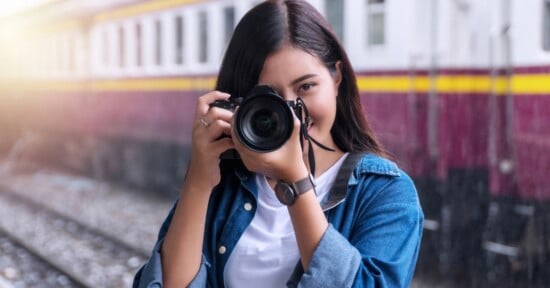 A young woman with long dark hair, wearing a blue denim jacket and white shirt, smiles while holding a camera up to her face at a train station, with train tracks and a train in the background.
